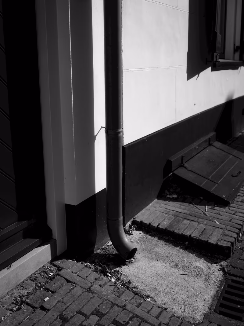 A metal downspout running down a rendered wall and turning outward, discharging onto brick paving immediately at the base of the building